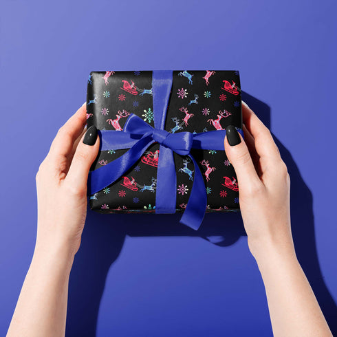 Christmas wrapping paper with a black background featuring colorful, stylized reindeer and Santa's sleigh designs. The reindeer and sleigh silhouettes are filled with skeletal patterns in shades of blue, pink, and red. Multicolored snowflakes in various sizes are scattered throughout. The paper has a whimsical, slightly gothic aesthetic that blends traditional holiday imagery with a unique, edgy twist.
